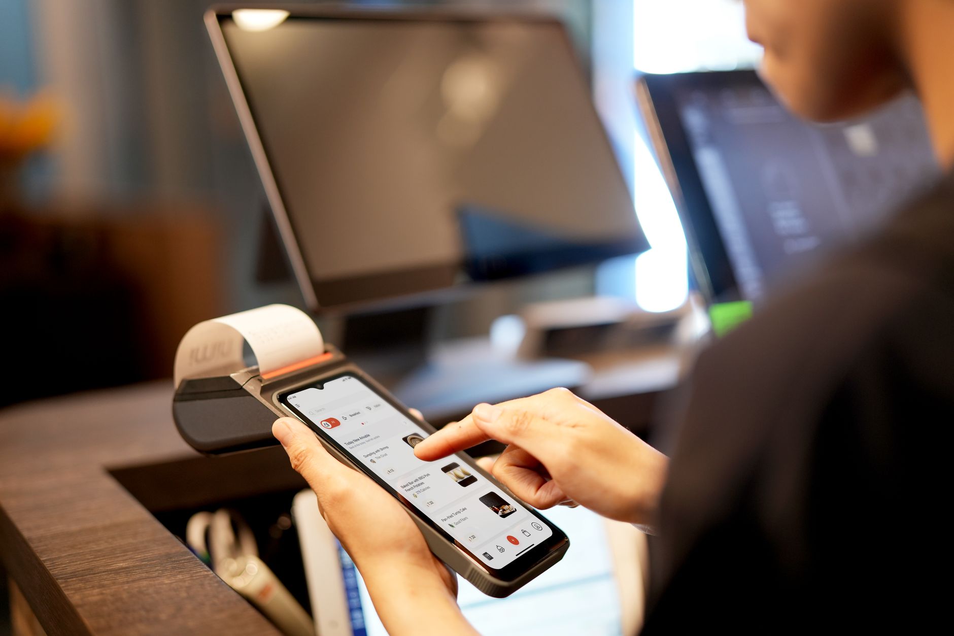 Une personne tient un smartphone devant un terminal de caisse, avec des écrans flous à l’arrière-plan.